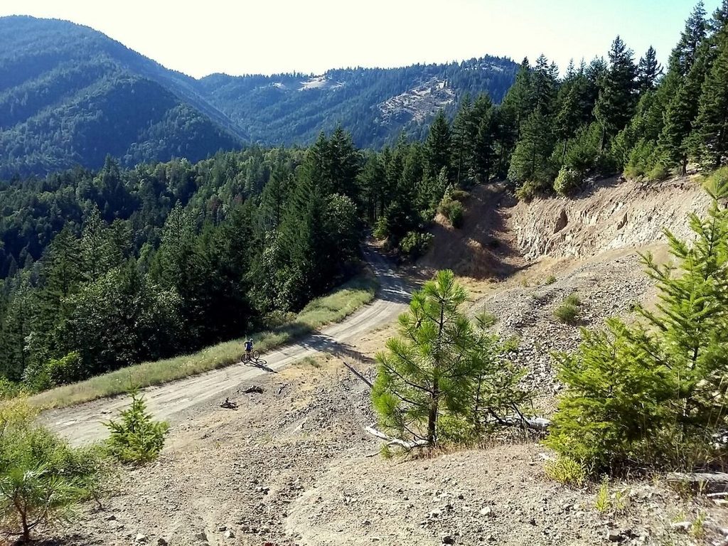A steep rocky descent drops to a dirt road, with forested ridges behind.