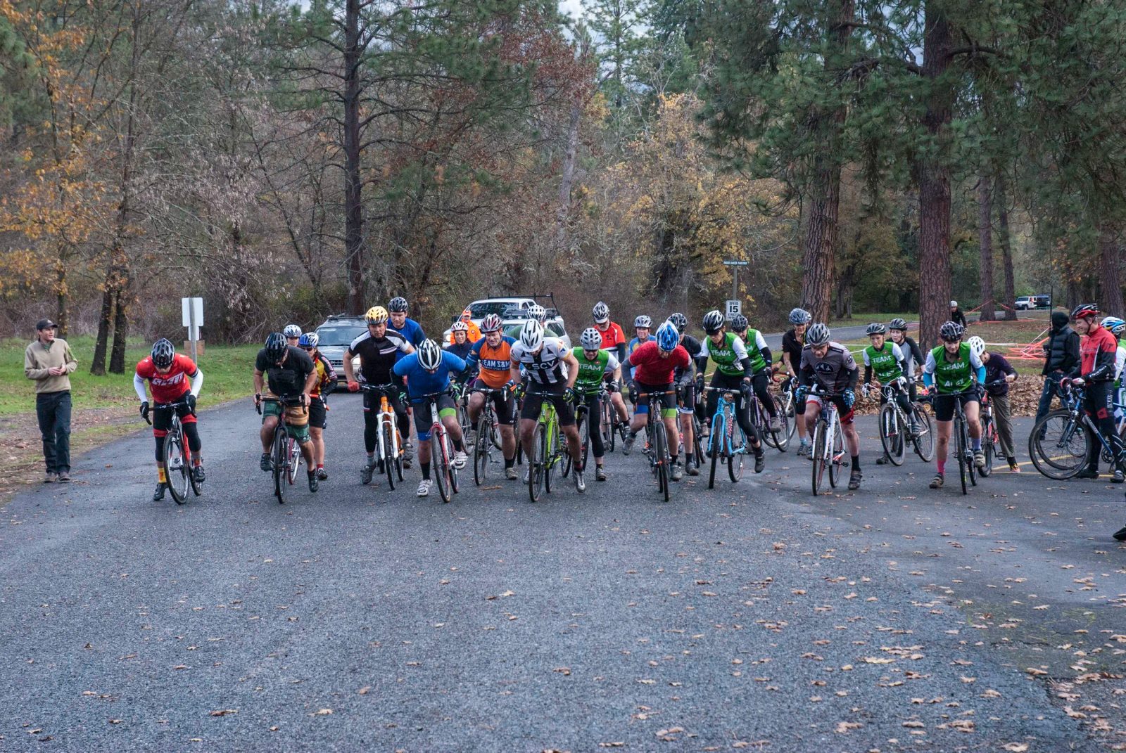 A mass start on the pavement at Tom Pearce Park, riders three abreast under the trees.