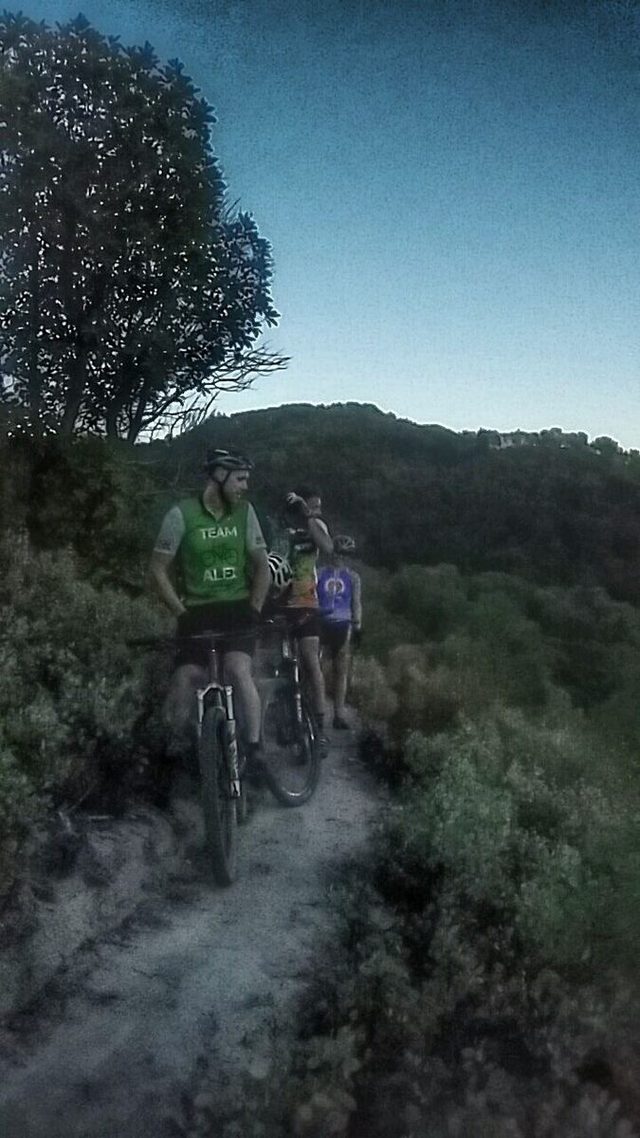 Two mountain bikers on a manzanita-lined singletrack at dusk.