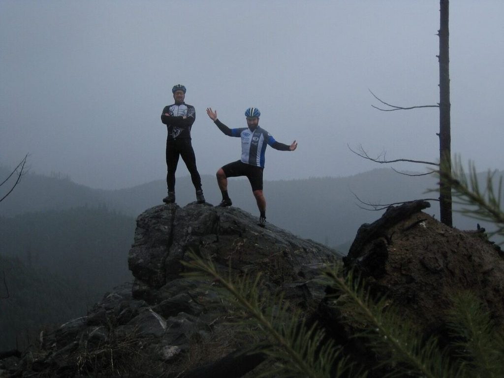 Two riders stand on a rock outcrop in the fog near the top of John's Peak.