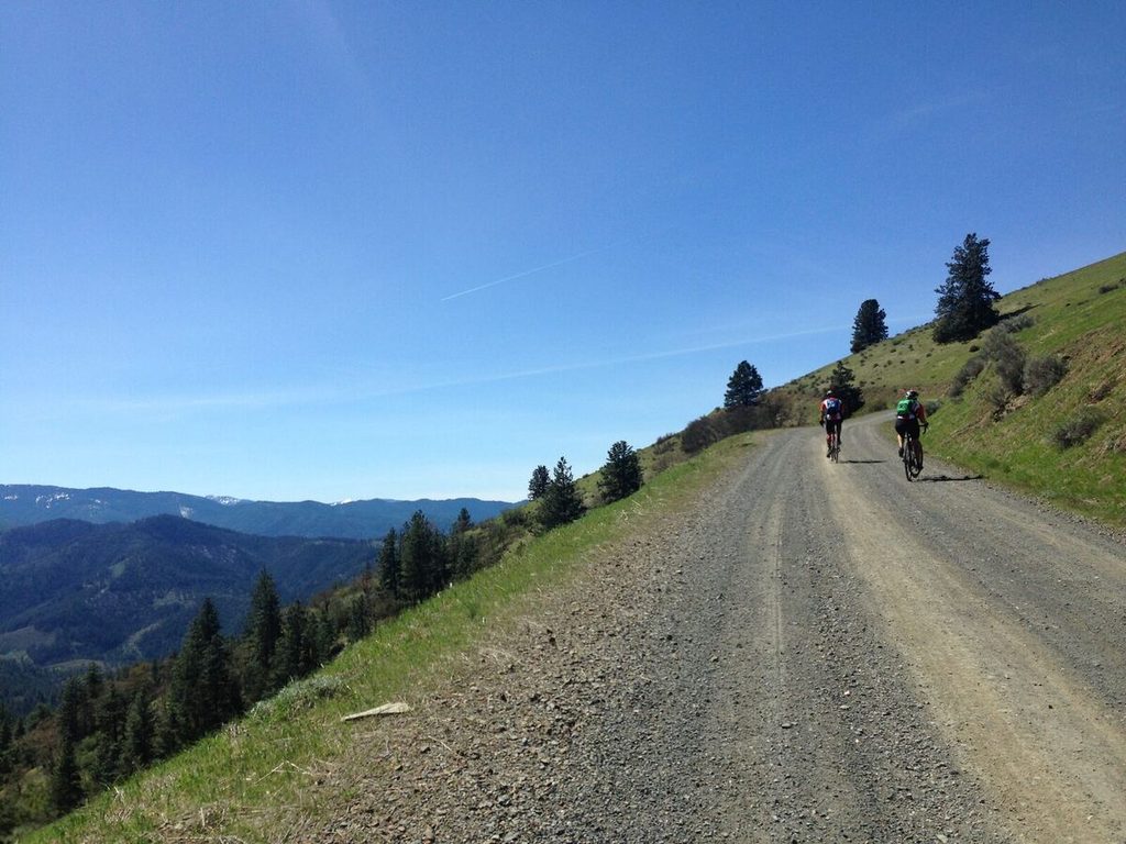 Two riders on a gravel ridge road on Anderson Butte above forested valleys.