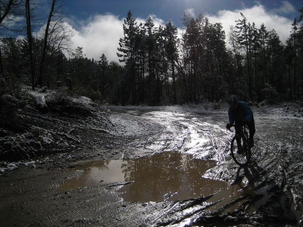 A rider on a muddy dirt road with puddles and patches of snow.